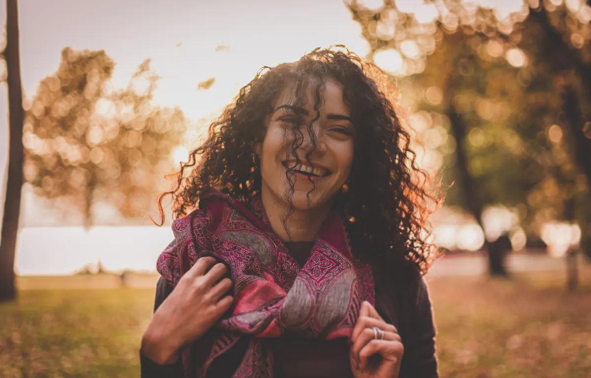 A woman with curly hair and a bright smile stands outdoors on a warm autumn day, soft golden-lit trees behind her.